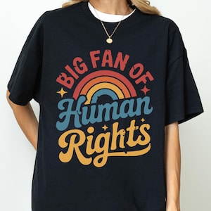 Könnte beinhalten: Schwarzes T-Shirt mit dem Text "BIG FAN OF Human Rights" in einem Retro-Regenbogen-Design. Das Shirt hat einen Rundhalsausschnitt und kurze Ärmel. Der Text ist im Vintage-Stil mit Regenbogen und Sternen gehalten.