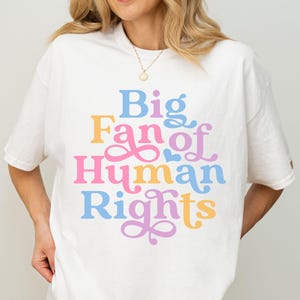 Puede incluir: Camiseta blanca con la frase "Big Fan of Human Rights" en letras coloridas y divertidas. La camiseta es de corte holgado y se combina con pantalones cortos negros. El estilo general es informal y cómodo.