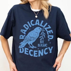 Può includere: T-shirt blu navy con grafica azzurra di un corvo con un ramoscello, circondato dalle parole "RADICALIZED" e "DECENCY", con "BY BASIC" sotto. La maglietta ha una vestibilità ampia.