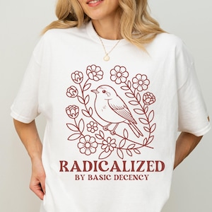 Op de afbeelding: Wit T-shirt met een bordeauxrode afbeelding van een vogel in een bloemenkrans. De tekst "RADICALIZED BY BASIC DECENCY" staat onder de afbeelding. De outfit wordt gecompleteerd met een zwarte short.