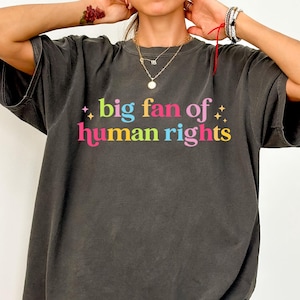 Könnte beinhalten: Dunkelgraues, übergroßes T-Shirt mit dem Text "big fan of human rights" in bunten Buchstaben. Das Shirt hat eine lockere Passform und kurze Ärmel. Der Text ist mit Sternen-Grafiken versehen.