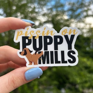 May include: A white sticker with black text that reads "pissin on PUPPY MILLS". A cartoon brown dog is shown lifting its leg to urinate on the text.