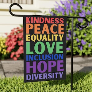 Może przedstawiać: Czarna flaga ogrodowa z napisem "KINDNESS, PEACE, EQUALITY, LOVE, INCLUSION, HOPE, DIVERSITY" w tęczowych kolorach. Flaga jest na czarnym metalowym stojaku w ogrodzie z kwiatami.
