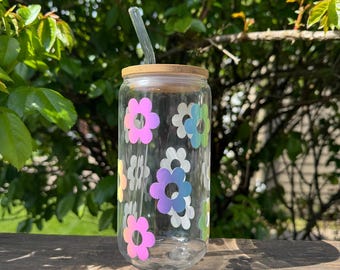 May include: A clear glass tumbler with a bamboo lid and a clear straw. The tumbler is decorated with colourful flower decals. The background is a blurred view of green foliage and a wooden surface.