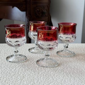 May include: Four clear glass cordial glasses with a red and gold metallic finish on the upper half. The lower half of each glass features a textured, patterned design. The glasses are displayed on a white surface.