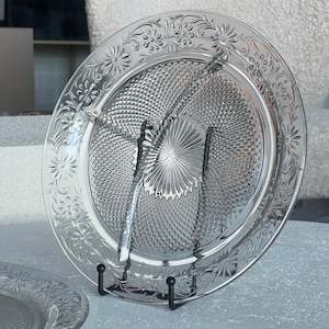 May include: A clear glass plate with a raised, diamond-shaped pattern and a floral design around the edge. The plate is on a black metal stand.