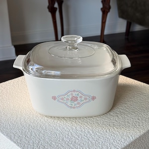 May include: A white casserole dish with a clear glass lid. The dish has a floral design in pink and blue. The dish has two handles and is sitting on a white surface.