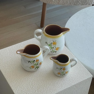 May include: Three white ceramic pitchers with brown rims and floral designs in yellow, green, and brown. The pitchers are of varying sizes and are arranged on a white surface.