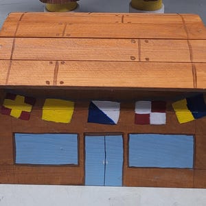 May include: A wooden box resembling a building, painted in shades of brown and orange. The front features blue windows and a blue door, with colorful flags above. The roof is made of wooden planks.