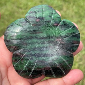 May include: A carved flower-shaped stone with green and black hues, accented with streaks of pink. The stone is held in a hand, showcasing its intricate details and natural patterns. The flower has five petals and carved lines.