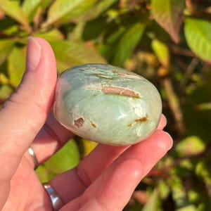 May include: A smooth, green and white polished gemstone with brown inclusions. The stone is shaped like an oval and is being held in a hand.