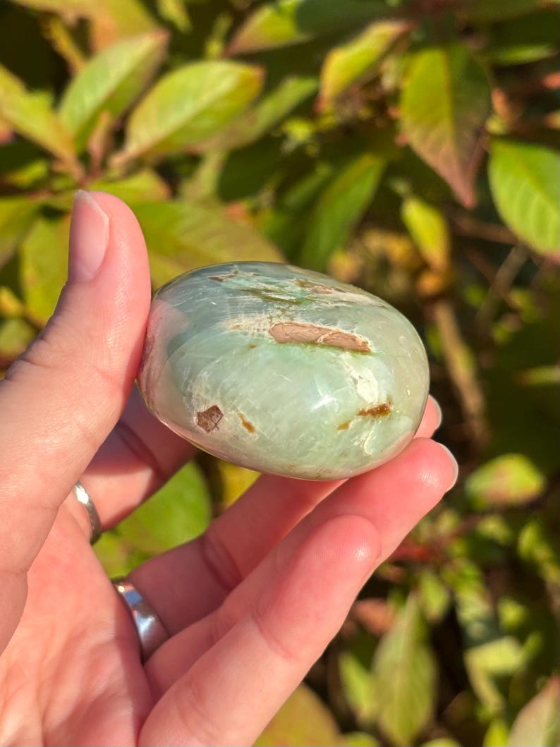May include: A smooth, green and white polished gemstone with brown inclusions. The stone is shaped like an oval and is being held in a hand.