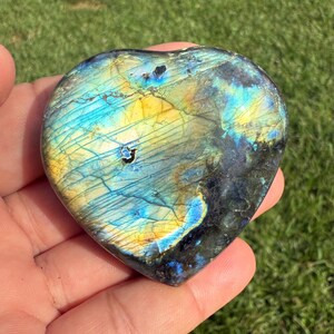 May include: A heart-shaped labradorite stone with a vibrant display of colors. The stone features a mix of blues, yellows, and blacks, with a shimmering, iridescent effect. The polished surface reveals natural patterns and textures, making it a unique decorative item.