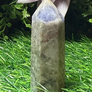 May include: A tall, polished crystal obelisk with a pointed top. The crystal has a gradient of colors, including gray, brown, and a purple hue at the top. It is set against a backdrop of green grass and purple flowers.