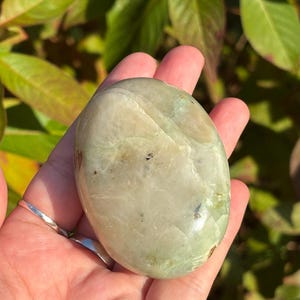 May include: A smooth, light green and white polished stone with a rounded shape. The stone is held in a hand.