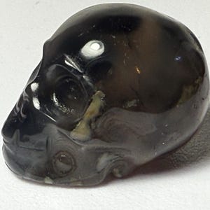 May include: A polished, dark-coloured skull carving. The skull is a mix of black, brown, and grey hues, with a glossy finish. The carving has detailed features, including eye sockets, a nose, and a jawline. The skull is a decorative item.