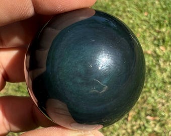 May include: A polished, dark teal obsidian sphere. The sphere has a glossy, reflective surface with swirling patterns and a subtle sheen. The stone is held between fingers against a backdrop of green grass.