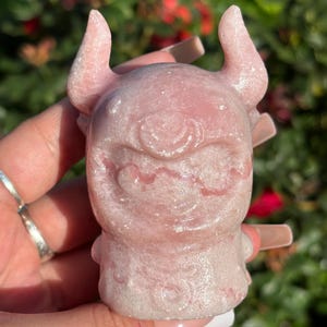 May include: A light pink, carved figurine of a cartoonish devil with horns, a single eye, and a wide grin. The figurine is held in a hand against a blurred background of green foliage and red flowers. The sculpture has a textured surface.