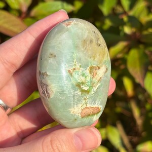 May include: A green and brown polished gemstone with a smooth, oval shape. The stone has a mottled pattern of green, brown, and white.