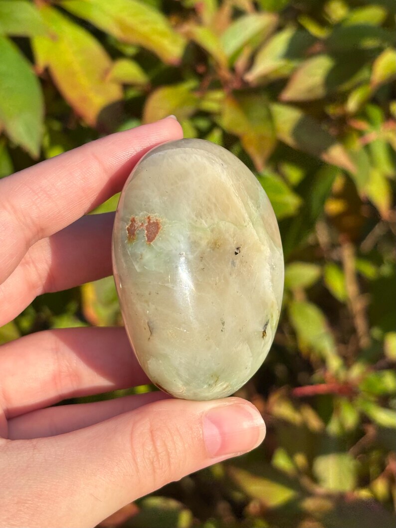May include: A smooth, oval-shaped green and white polished stone with brown and black inclusions. The stone is held in a hand.
