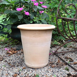 Peut inclure: Un grand pot de fleurs en terre cuite non vernissée avec un large bord. Le pot est cylindrique et repose sur un lit de petits graviers. En arrière-plan, des plantes vertes, une chaise en fer forgé et un mur de briques créent un cadre de jardin.