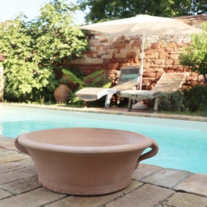Op de afbeelding: Een terracotta plantenbak met twee handvatten staat op een betegeld terras naast een zwembad. Het zwembad is omgeven door weelderig groen en een bakstenen muur met twee ligstoelen.