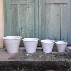 Puede incluir: Cuatro macetas de cerámica blanca de diferentes tamaños, dispuestas en fila. Las macetas tienen una superficie ligeramente texturizada y un diseño sencillo. El fondo es una puerta de madera desgastada de color turquesa.