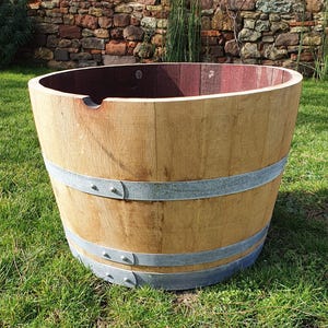 Pode incluir: Um grande plantador de barril de madeira com um acabamento de madeira natural. O barril é mantido unido por duas faixas de metal e assenta sobre relva verde. O interior do barril é de uma cor castanho-avermelhada mais escura.