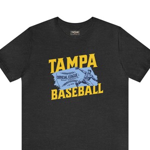 Pode incluir: Uma camiseta cinza escura com um gráfico vintage de um jogador de beisebol e o texto "TAMPA OFFICIAL LEAGUE FULLY GUARANTEED BASEBALL".