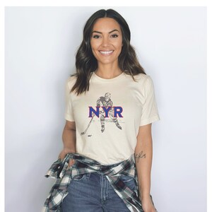 May include: A woman wearing a cream-colored t-shirt with a vintage hockey player graphic and the text "NYR" in blue and red. She is wearing a plaid shirt tied around her waist and blue jeans.