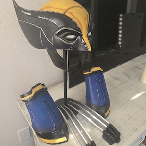 Op de afbeelding: Een zwart-gele helm met puntige zijkanten en witte ooglenzen, vergezeld door twee blauw-gele onderarmbeschermers en een set van drie lange, zilverkleurige metalen klauwen, alles tentoongesteld op een zwarte standaard met een centrale steun.