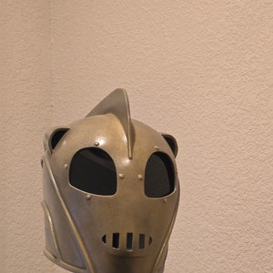 Puede incluir: Un casco de color dorado con una parte superior puntiaguda y una máscara facial con dos grandes agujeros para los ojos y una fila de pequeños agujeros para la boca. El casco está en un soporte negro.
