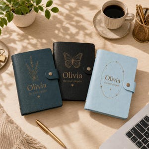 May include: Three personalised journals in dark blue, black, and light blue with gold foil designs. Each features the name Olivia, the text the next chapter, and a snap closure. The dark blue has a floral sprig, the black has a butterfly, and the light blue has an oval frame.