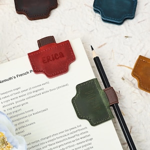 May include: Leather page markers in shades of red, green, and brown. The red marker, embossed with "Erica", is on a page. A pencil is placed through a green marker. The page displays a recipe. Measurements are in grams and cm.