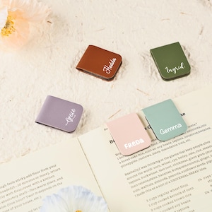 Puede incluir: Un conjunto de cinco marcapáginas de cuero personalizados en marrón, lavanda, verde oliva, rosa y turquesa. Cada marcapáginas presenta un nombre diferente en escritura blanca: Hedda, Grace, Ingrid, Freda y Gemma. Los marcapáginas están sobre un libro abierto.