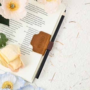 Op de afbeelding: Een bruine leren pennenhouder met de naam "Olivia" erin geëmbosseerd, bevestigd aan een notitieboek met een zwarte potlood. Het notitieboek is open op een receptpagina. De afbeelding is versierd met bloemen.