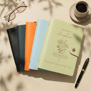 Pode incluir: Uma coleção de diários personalizados em várias cores: preto, verde-azulado, laranja, azul claro e verde claro. O diário verde claro apresenta o texto "THE NEXT chapter" e o nome "NICHOLAS". Uma chávena de café, óculos e uma caneta também estão presentes.