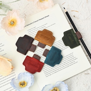 Könnte beinhalten: Lesezeichen aus Leder in verschiedenen Farben, jeweils mit einem eingeprägten Namen. Die Farben sind Braun, Beige, Rot, Blau und Grün. Ein Bleistift liegt auf dem offenen Buch.