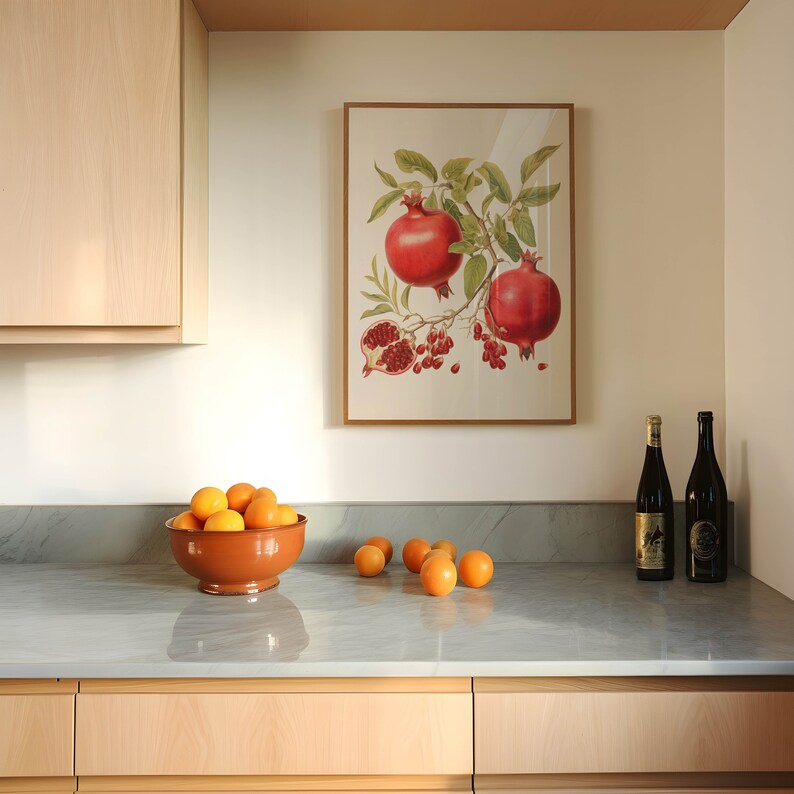 May include: A framed botanical print of a pomegranate branch with red fruit and green leaves hangs on a white wall above a kitchen counter with a bowl of oranges and two bottles of wine.