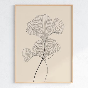 Op de afbeelding: Een minimalistische lijnkunstprint van een ginkgo-blad in zwart op een beige achtergrond. De print is ingelijst in een licht houten lijst.