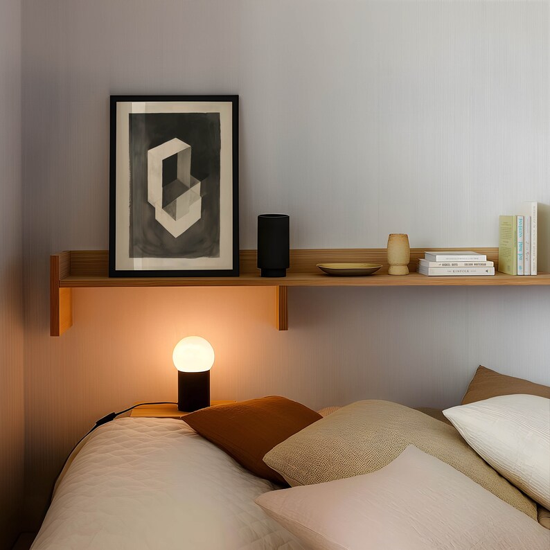 May include: A bedroom with a wooden shelf with a framed print, a speaker, a vase, and books. A bedside table with a lamp and a white bed with multiple pillows.