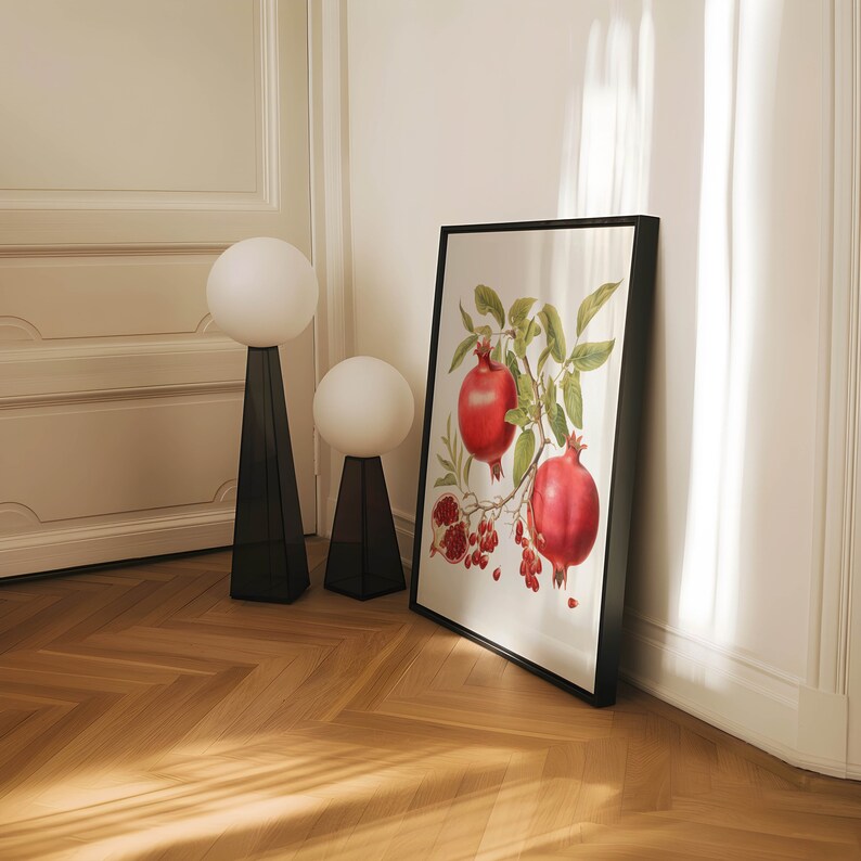 May include: A black framed print of a botanical illustration of a pomegranate branch with red pomegranates and green leaves. The print is hanging on a white wall next to two white globe lamps with black bases.