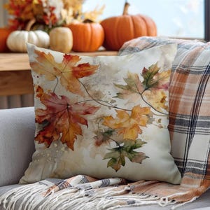 Peut inclure: Un coussin carré orné d'une impression aquarelle de feuilles d'érable d'automne dans des tons orange, rouge, jaune et vert sur un fond crème. Le motif comprend des tiges brunes délicates et de légères éclaboussures de peinture.