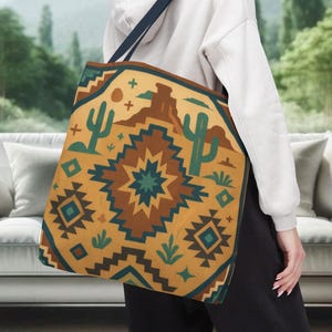 May include: A tan tote bag with a Southwestern design featuring cacti, mountains, and geometric patterns in shades of brown, teal, and gold. The bag has a navy blue strap and is carried over the shoulder.