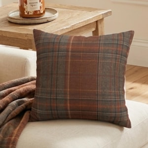 May include: A square throw cushion featuring a plaid pattern in shades of brown, grey, and orange. The cushion is placed on a light-coloured sofa next to a folded matching plaid blanket, with a candle in a glass jar visible in the background.
