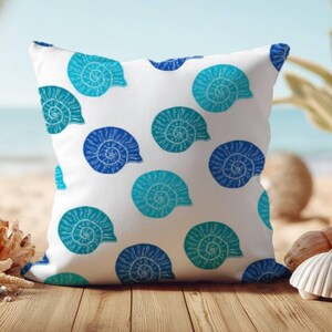 Op de afbeelding: Een vierkant sierkussen met een witte achtergrond voorzien van een herhalend patroon van spiraalvormige schelpen in diverse blauwtinten, waaronder licht turquoise, medium cyaan en diep marineblauw, verspreid over de voorzijde.