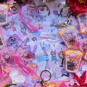 Könnte beinhalten: Eine Sammlung von Acryl-Schlüsselanhängern mit Zeichentrickfiguren, darunter Gestalten in rosa und lila Outfits sowie Figuren in Behältern mit goldenem Glitzer. Die Schlüsselanhänger haben goldfarbene Metallringe und liegen auf einer gemusterten Fläche.
