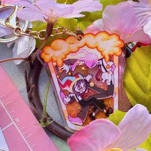 May include: A clear acrylic shaker keychain featuring a cartoon character with a jester hat and a pink rabbit character inside a container filled with orange glitter. The keychain has a gold-tone metal ring and is shown next to a pink ruler.