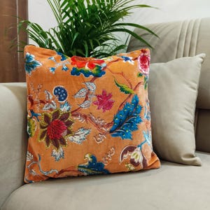 May include: An orange cushion featuring a floral design in blue, red, and green. The cushion is placed on a beige sofa, with a second beige cushion and a green plant visible in the background.