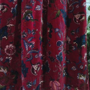 May include: A close-up of a red fabric with a floral pattern. The design includes blue, green, and white flowers and leaves. The fabric is draped, with folds and shadows, creating a textured appearance. The overall theme is a vintage or botanical textile.
